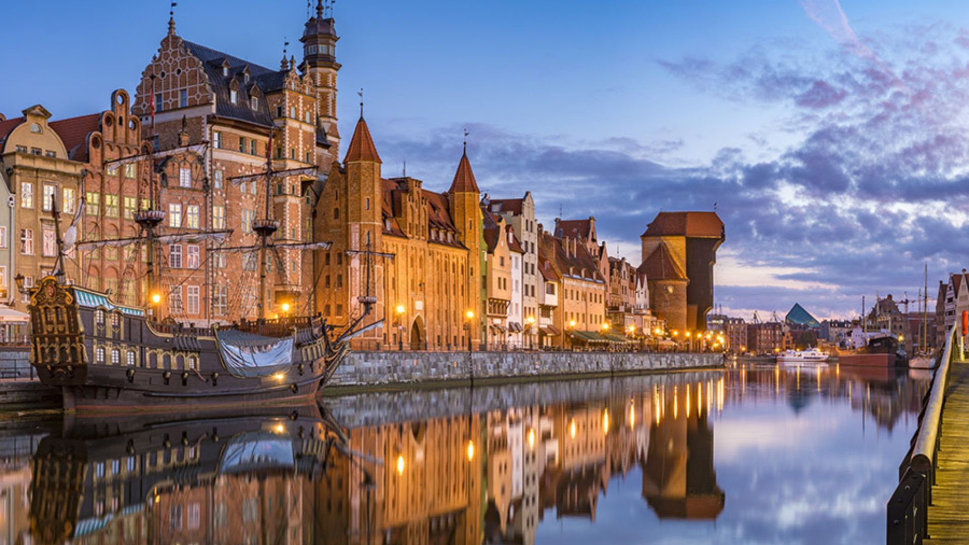 Un galion d'époque est amarré le long d'un canal de la ville au crépuscule, avec des bâtiments historiques illuminés se reflétant dans l'eau calme.