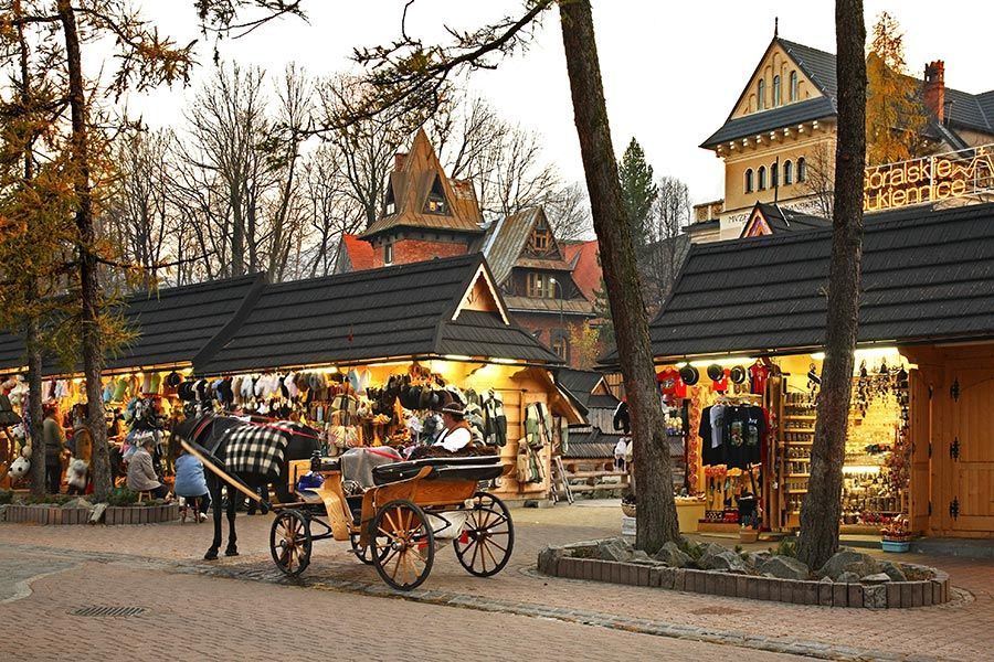 A horse-drawn carriage is parked on a brick street beside illuminated wooden market stalls, with rustic buildings and trees in the background.