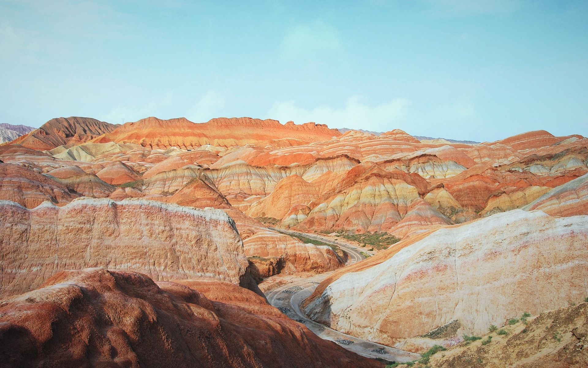 Un vasto paesaggio di montagne colorate e stratificate con sfumature di arancione, rosso e giallo sotto un cielo azzurro chiaro.