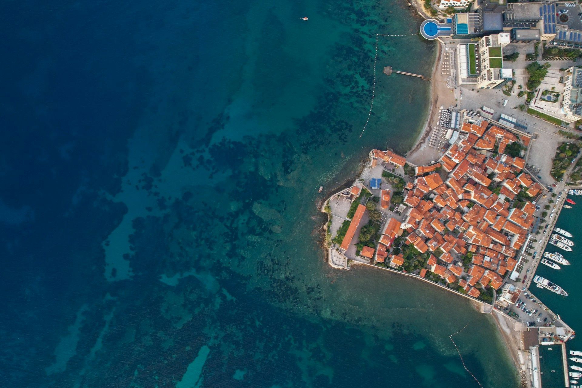 Una veduta aerea, dall'alto, di una città costiera con tetti in tegole arancioni che si affaccia su acque turchesi e limpide, accanto a un porto turistico.