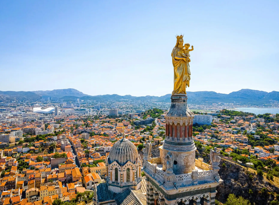 Una statua dorata di madre e figlio sulla guglia di una cattedrale domina una vasta città costiera con tetti in terracotta.