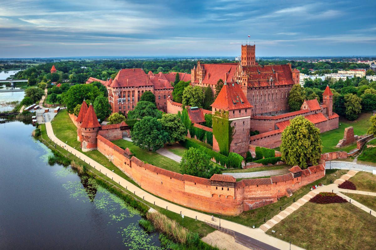 Vue aérienne d'un grand château médiéval en briques rouges avec des tours, entouré d'un mur fortifié et d'une rivière, au milieu d'arbres verdoyants.