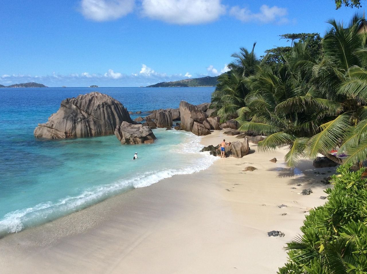 Ein erhöhter Blick auf einen tropischen Strand mit weißem Sand, türkisblauem Wasser, großen Granitfelsen und Palmen unter sonnigem Himmel.
