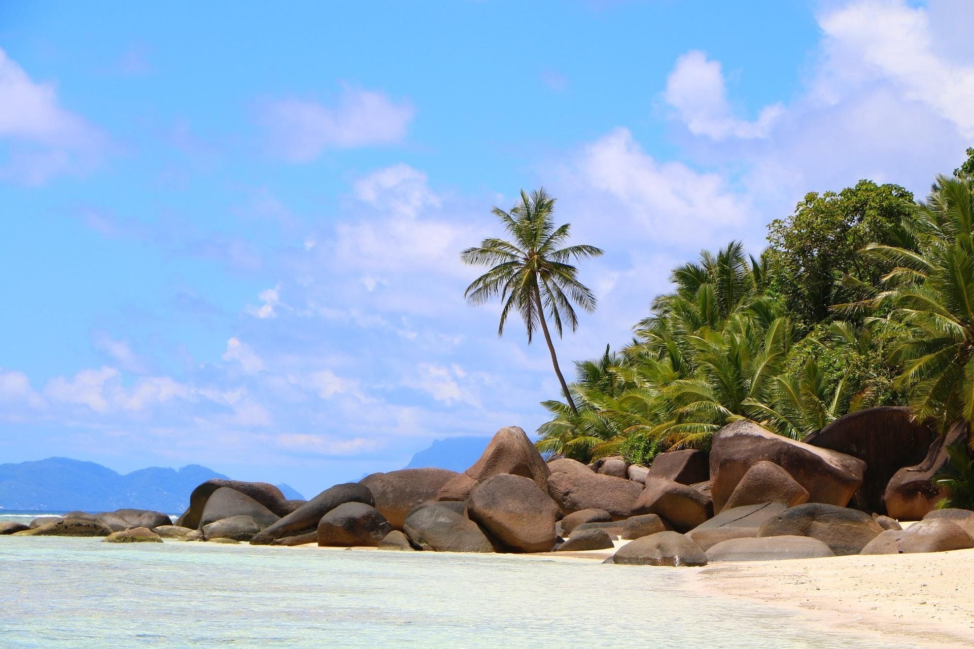 Ein tropischer Sandstrand mit großen, glatten Felsen und üppigen Palmen am klaren Wasser, unter blauem Himmel mit Wolken.