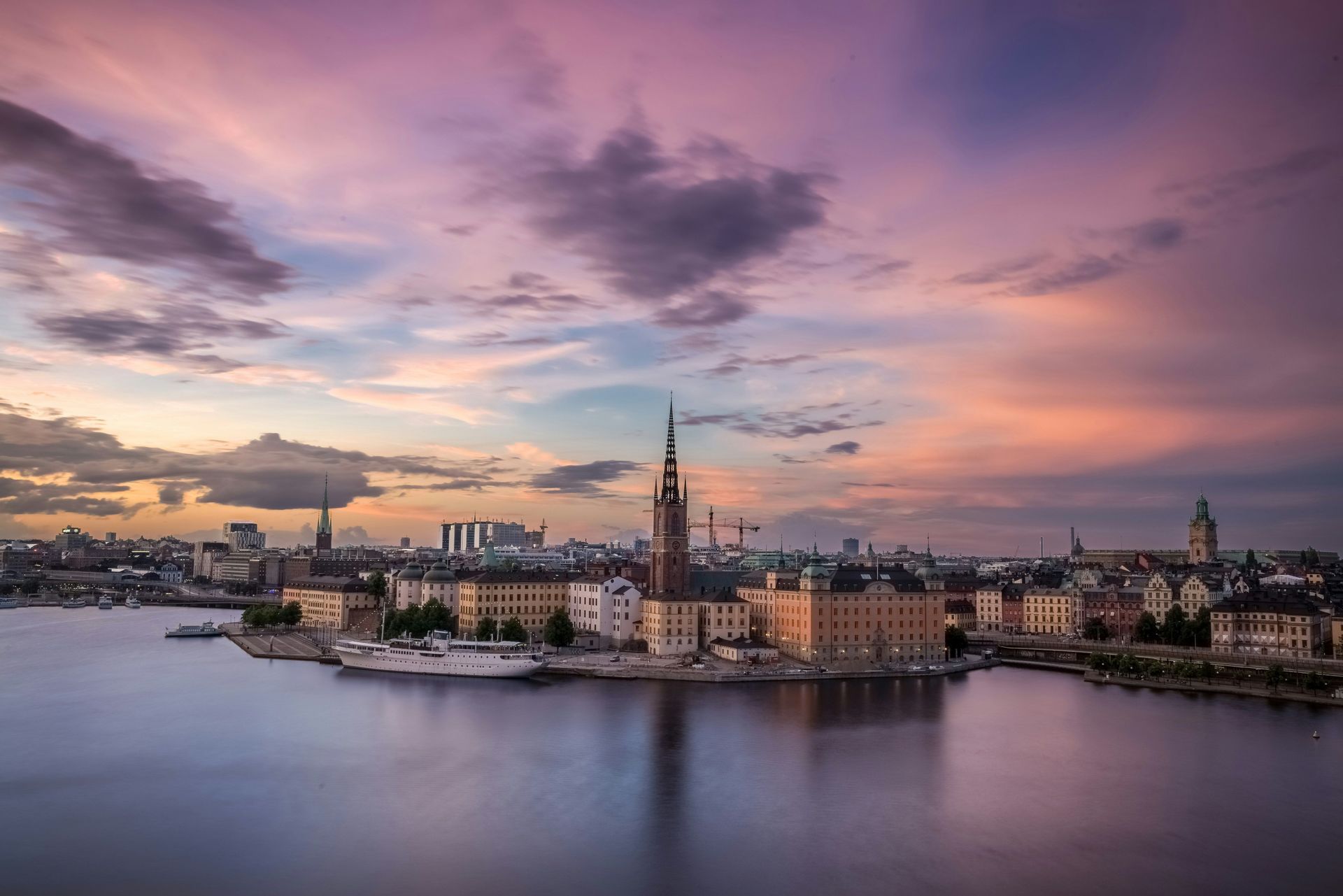 Uno skyline cittadino lungo un corso d'acqua, con un'alta guglia di chiesa, è illuminato da un vibrante tramonto viola e rosa.