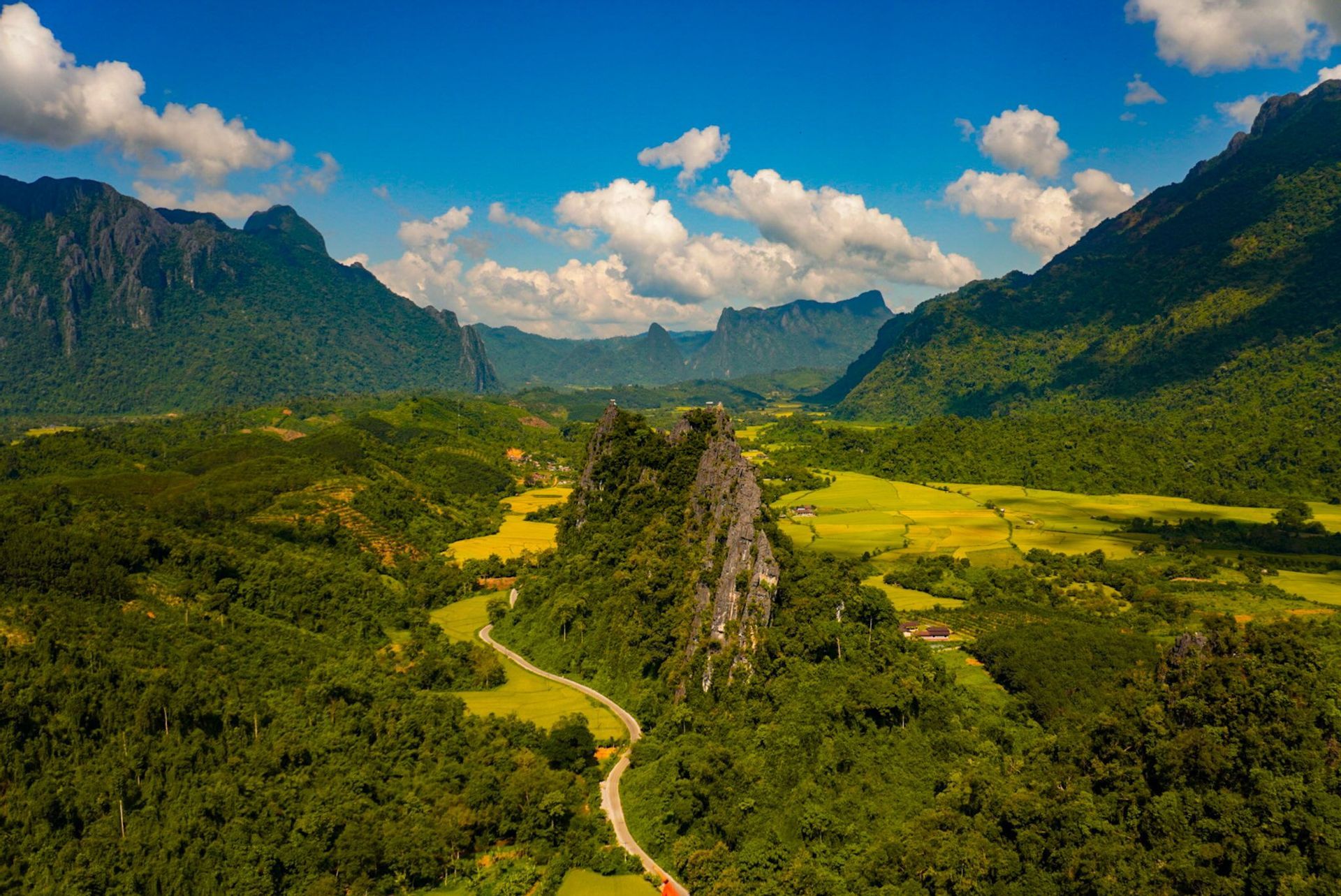 Une vue aérienne d'une route sinueuse à travers une vallée verdoyante avec des rizières, entourée de majestueuses montagnes calcaires sous un ciel bleu.