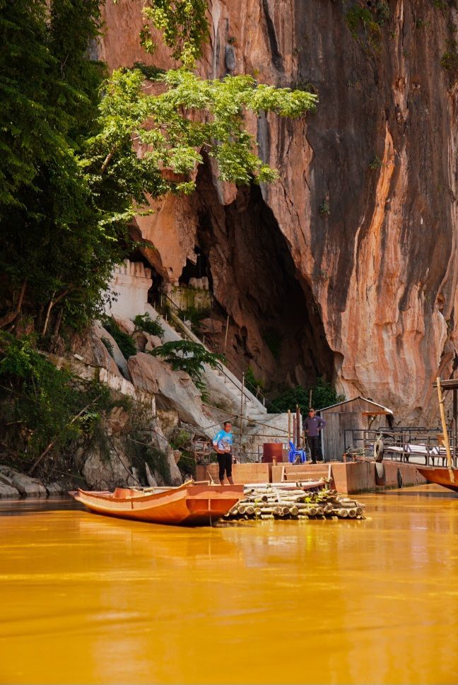Une pirogue à queue longue est amarrée à un ponton en bois sur une rivière boueuse, devant une grande falaise avec une entrée de grotte.