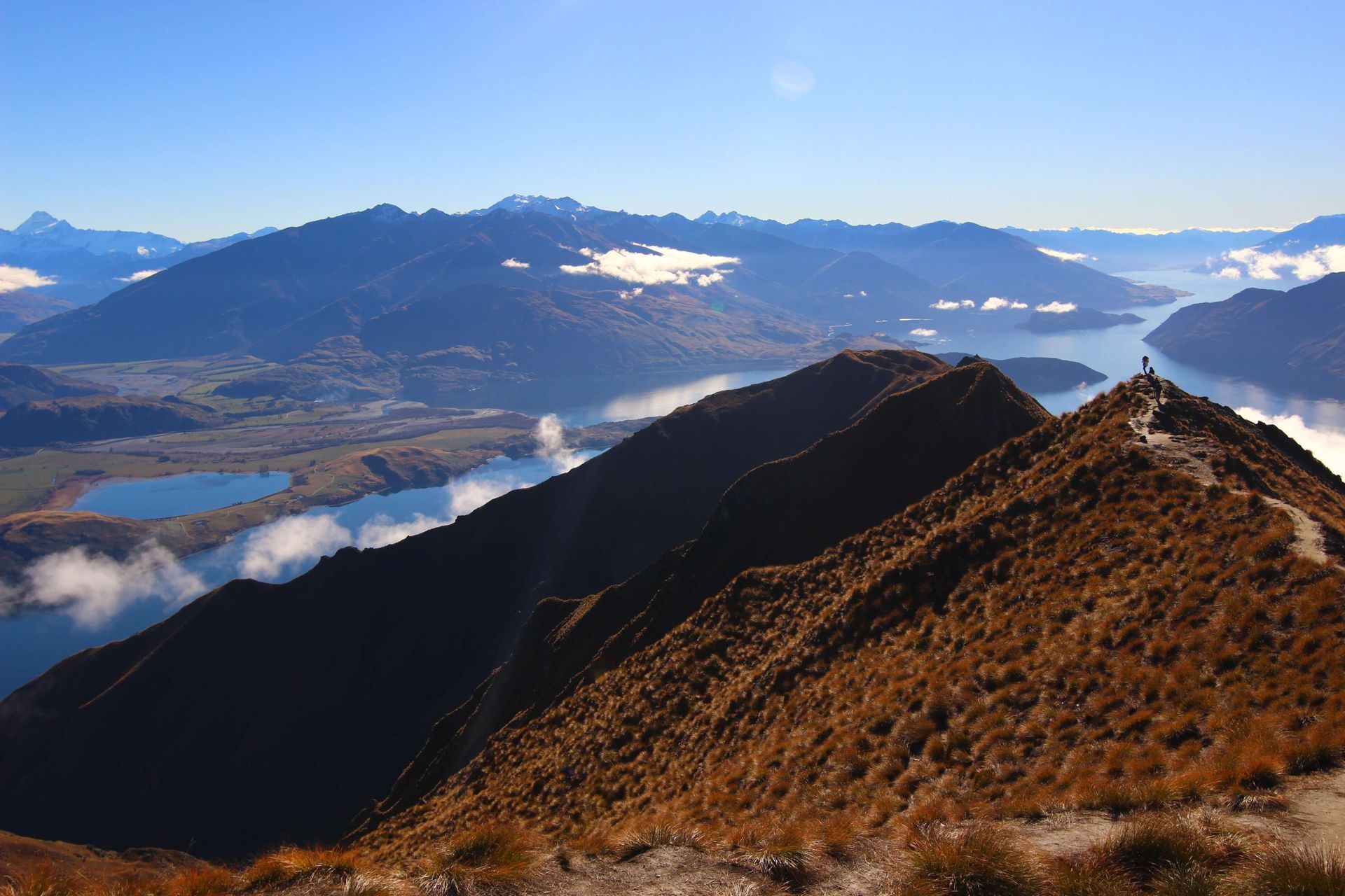 A grassy mountain ridge leads to a peak with two hikers overlooking a vast landscape of lakes and mountains under a blue sky.