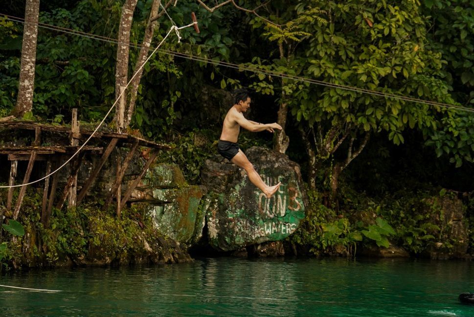Un homme torse nu en short noir saute dans une rivière verte depuis un rocher dans une jungle luxuriante.