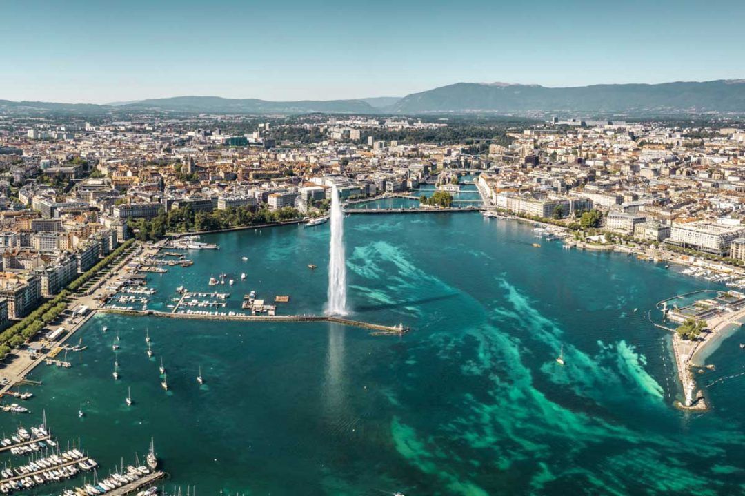An aerial view of a large water fountain spraying from a turquoise lake, with a dense cityscape and marina along the shore.