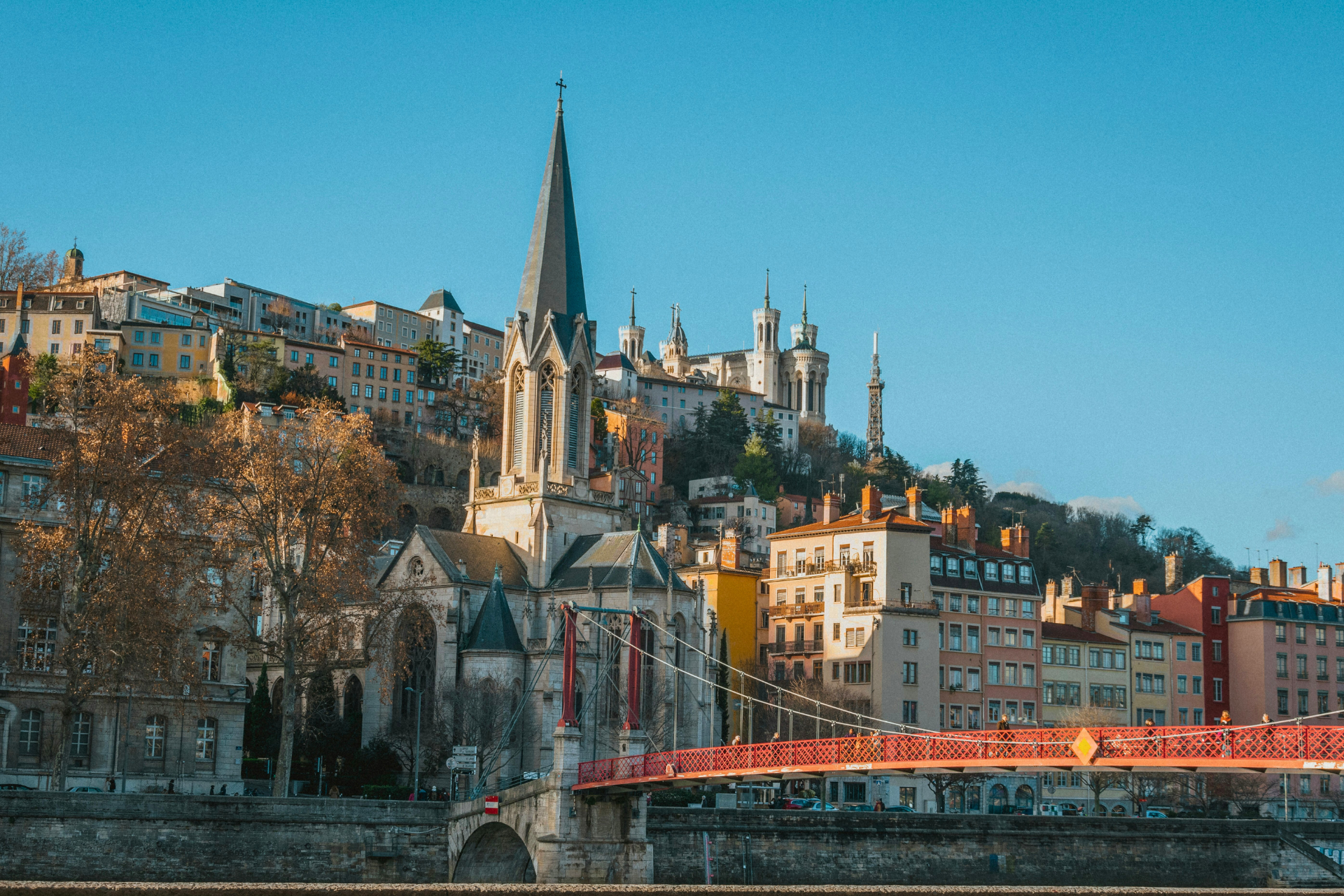 Un pont rouge enjambe une rivière devant une église gothique et une ville à flanc de colline, sous un ciel bleu clair.