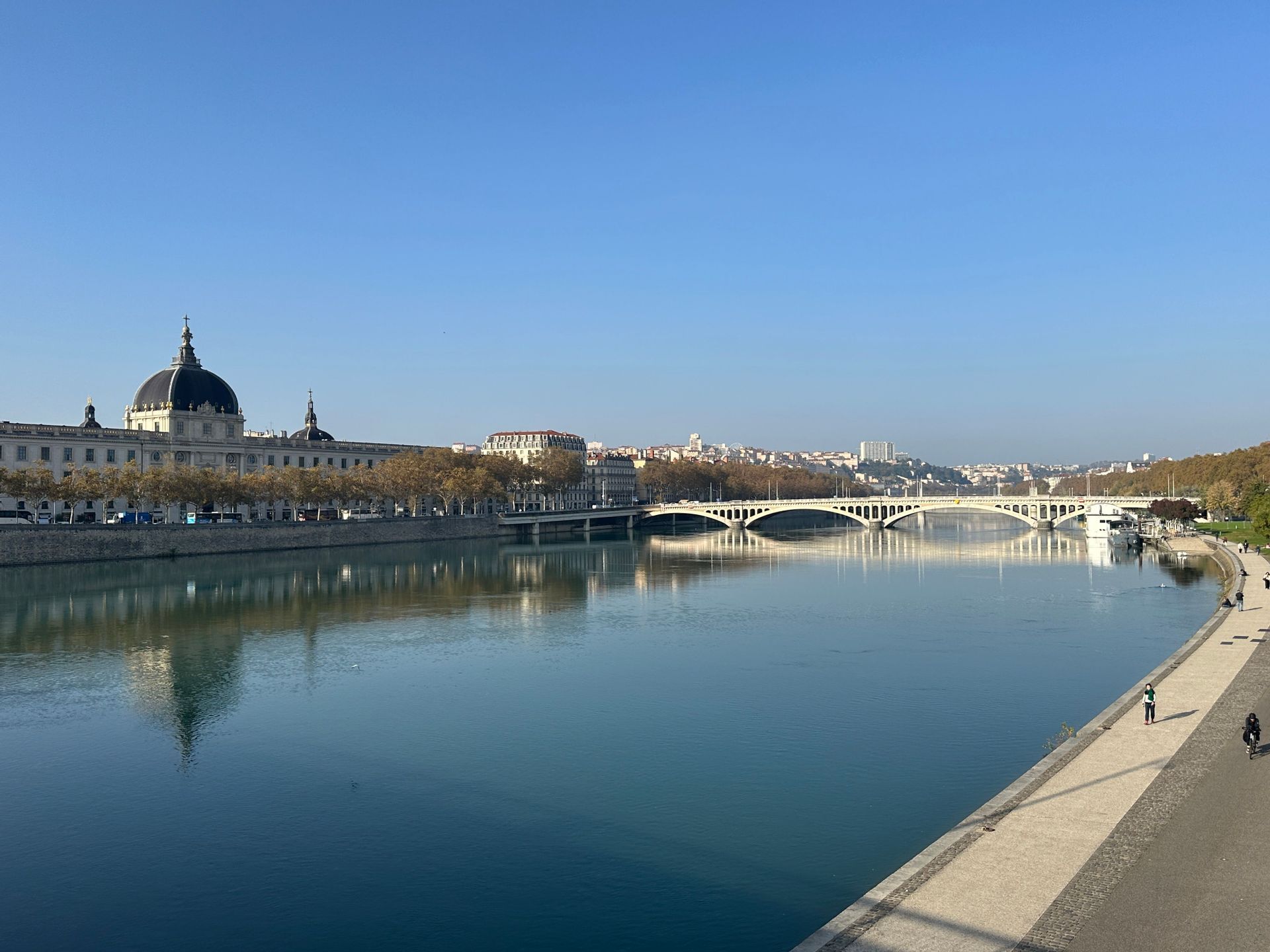 Une large rivière traverse une ville par une journée ensoleillée, avec un bâtiment en dôme sur une rive et un pont arqué au loin.
