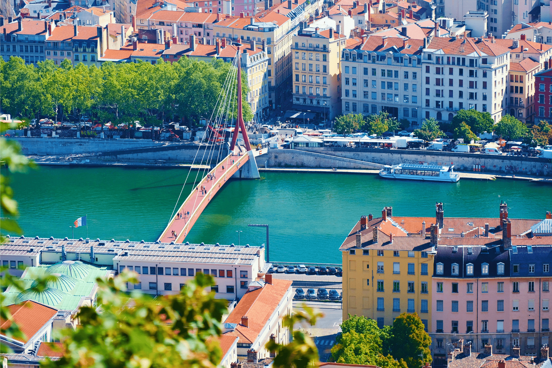 Vogelperspektive auf eine Stadtlandschaft mit einer roten Fußgängerbrücke über einem türkisfarbenen Fluss, Gebäuden und Bäumen entlang der Ufer.
