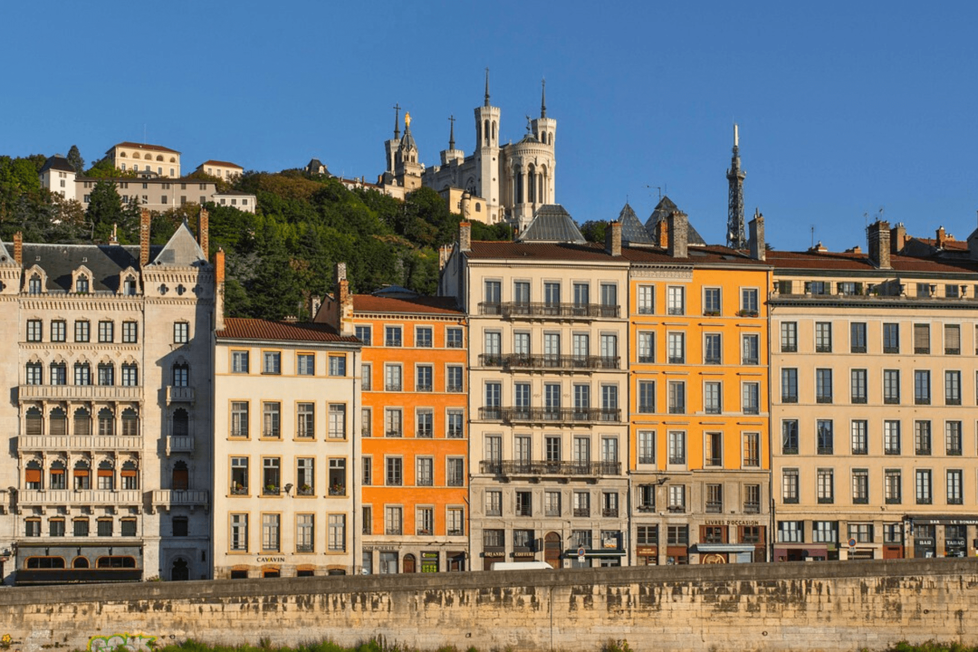 Des bâtiments colorés bordent une rivière, avec une cathédrale blanche sur une colline verdoyante en arrière-plan, sous un ciel bleu clair.