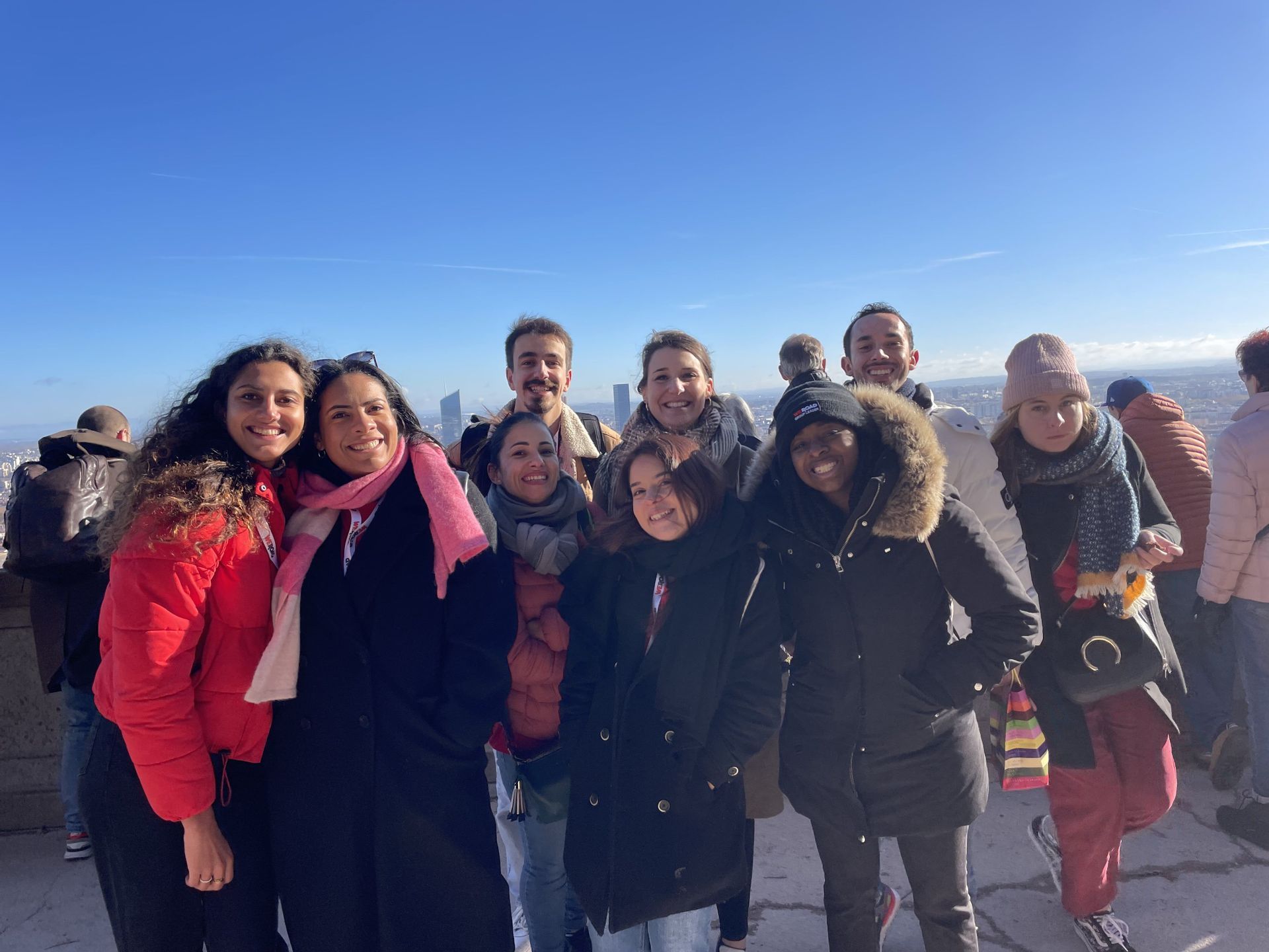 Un groupe WeRoad de neuf personnes souriant pour une photo, vêtues de manteaux d'hiver depuis un point de vue de la ville sous un ciel bleu clair.