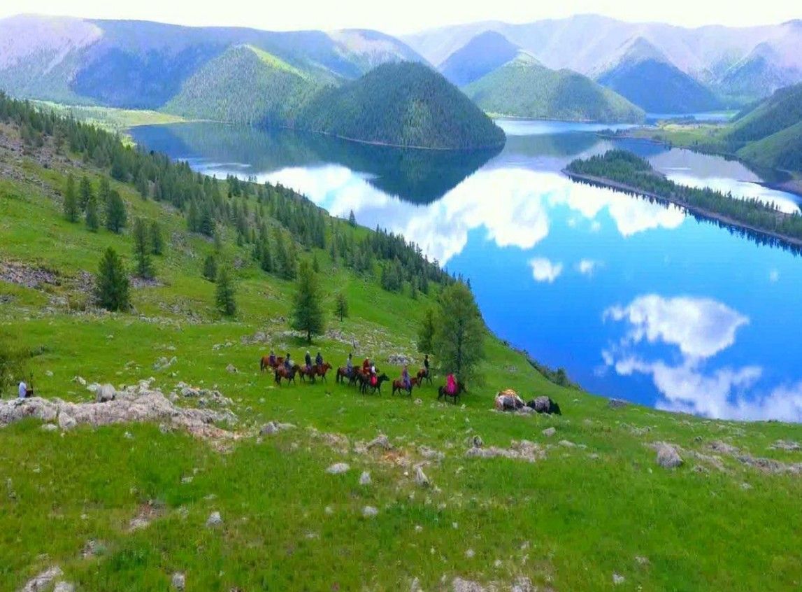Eine WeRoad Gruppenreise zu Pferd ruht auf einem grünen Hügel mit Blick auf einen ruhigen See, der den Himmel und die Berge widerspiegelt.