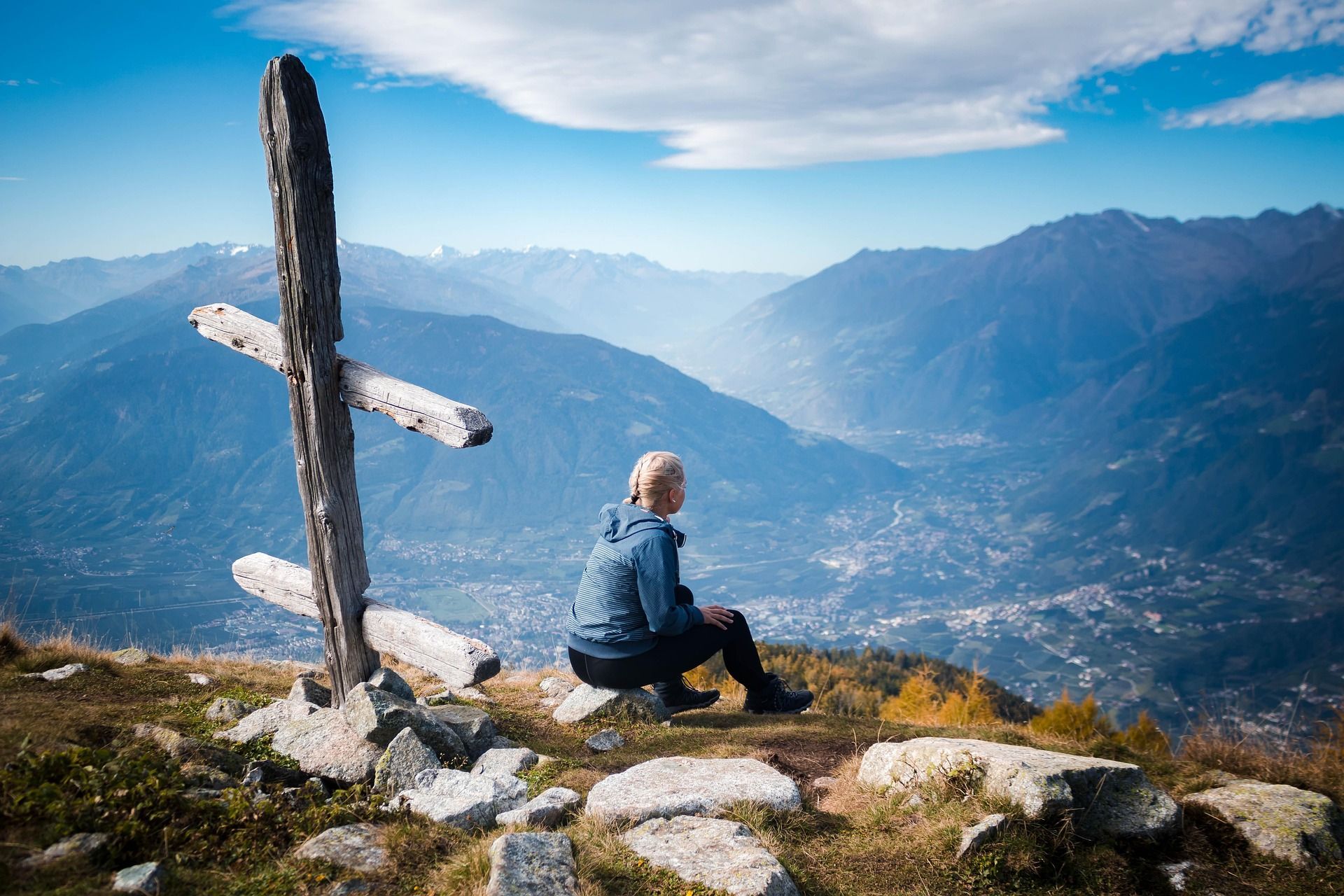 Una persona con trenzas rubias se sienta en un afloramiento rocoso junto a una cruz de madera, con vistas a un amplio valle de montaña.