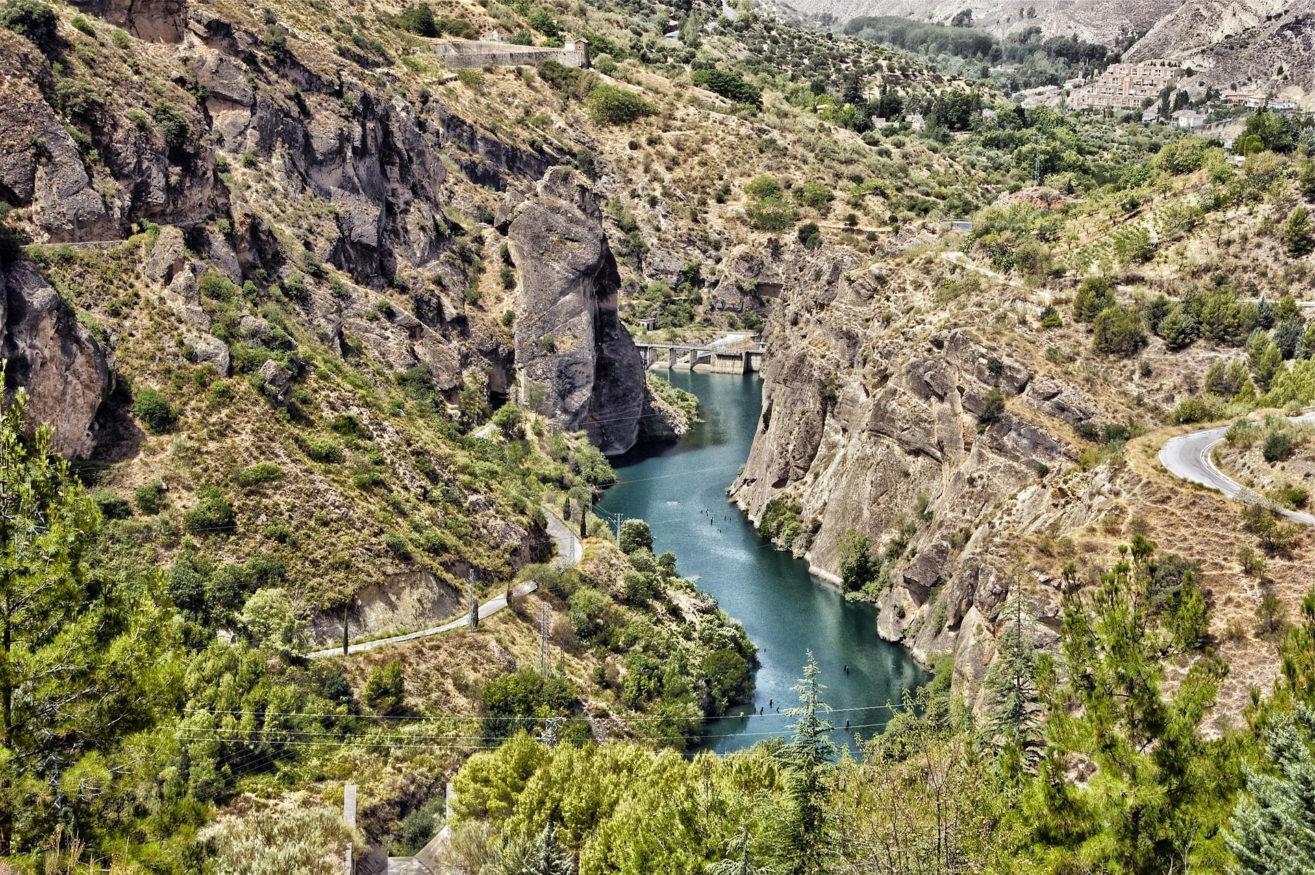 Vista aérea de un río turquesa serpenteando por un cañón rocoso y escarpado, con laderas áridas cubiertas de vegetación y una presa distante.