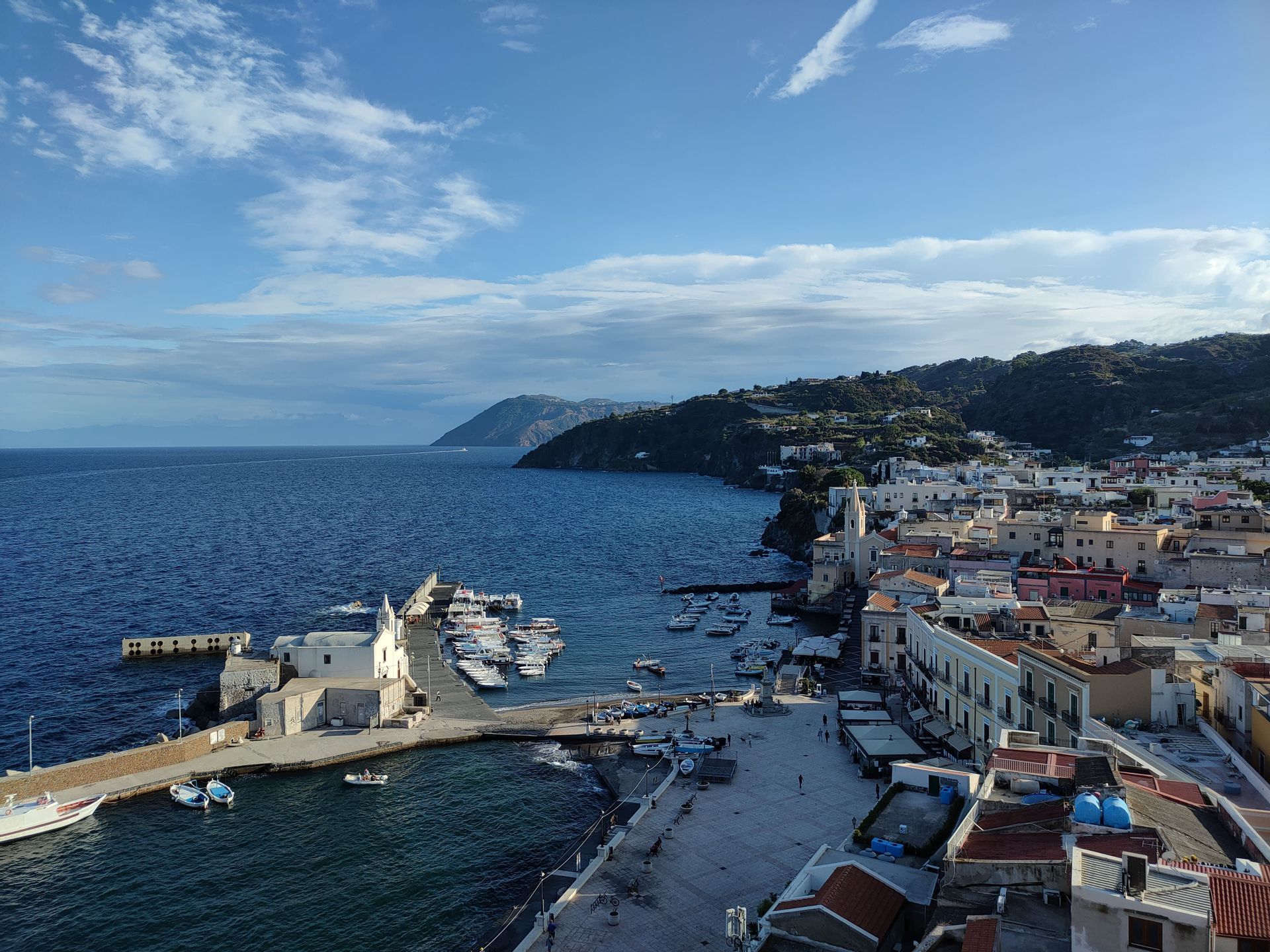 Vista aerea di una colorata cittadina costiera e del suo porto pieno di barche, adagiata su un litorale collinare sotto un cielo azzurro.