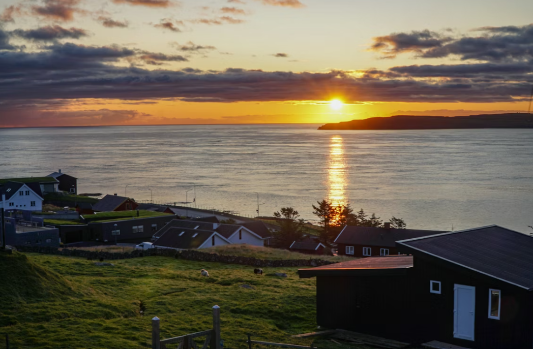Un petit village côtier sur une colline verdoyante avec le soleil se couchant sur la mer calme, projetant un reflet doré sur l'eau.
