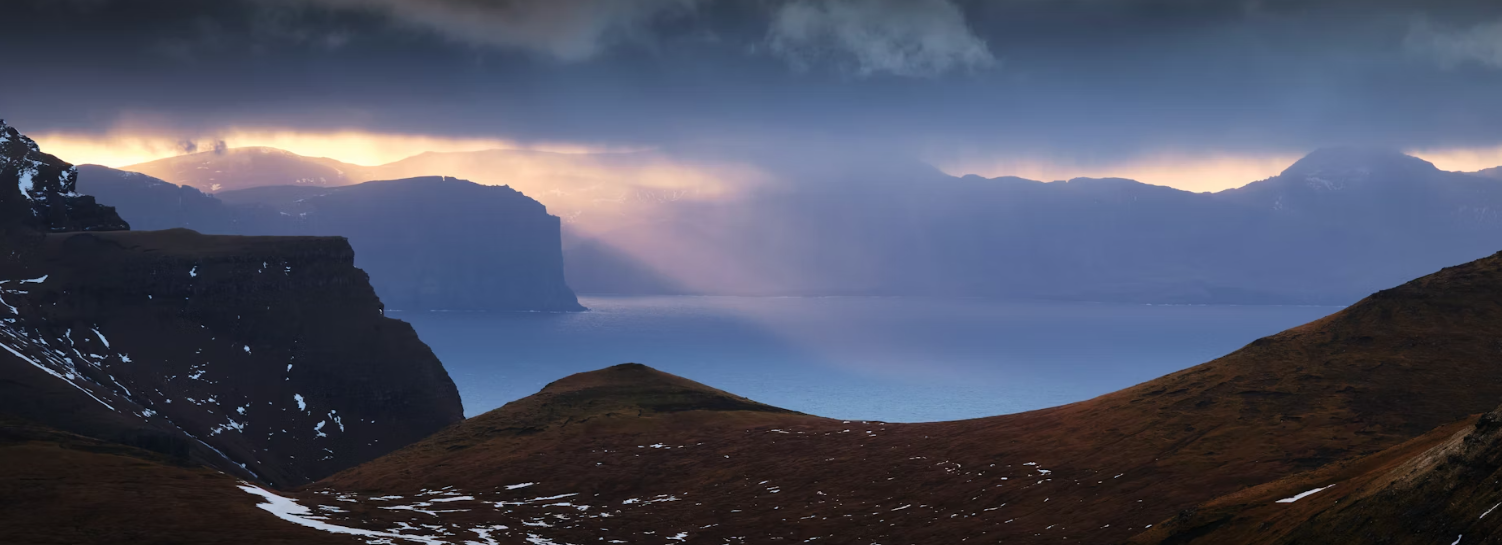 Un rayon de soleil perce les nuages sombres au-dessus d'un fjord paisible, avec des montagnes escarpées au loin et des collines verdoyantes au premier plan.
