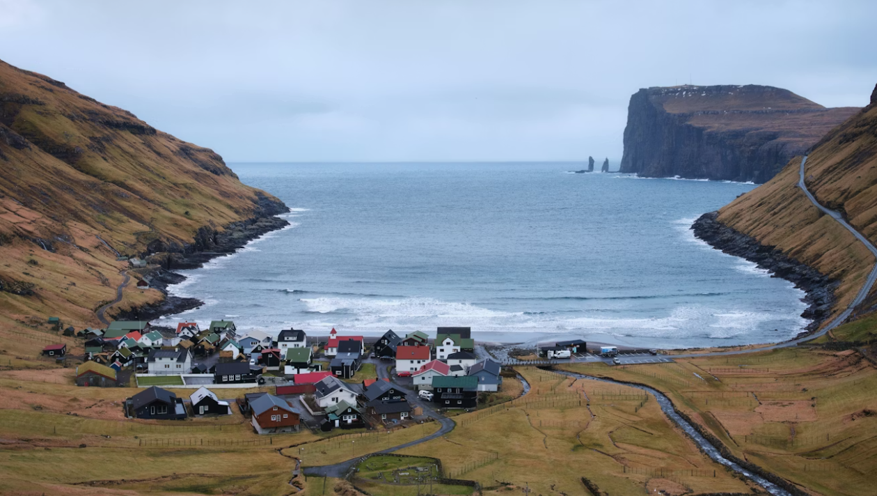 Vue aérienne d'un petit village aux maisons colorées, niché au bord d'une baie, entouré de collines verdoyantes et escarpées sous un ciel nuageux.