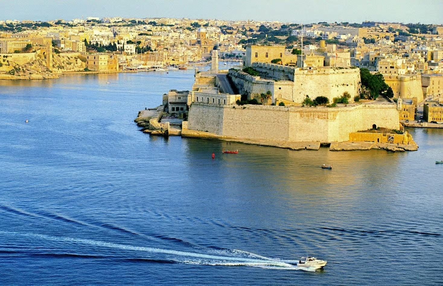 A white motorboat speeds across blue water, creating a wake, with a large stone fortress and coastal city in the background.