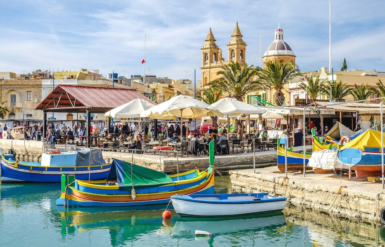 Colorful boats are docked in a harbor next to bustling waterfront cafes, with a historic town and church in the background.