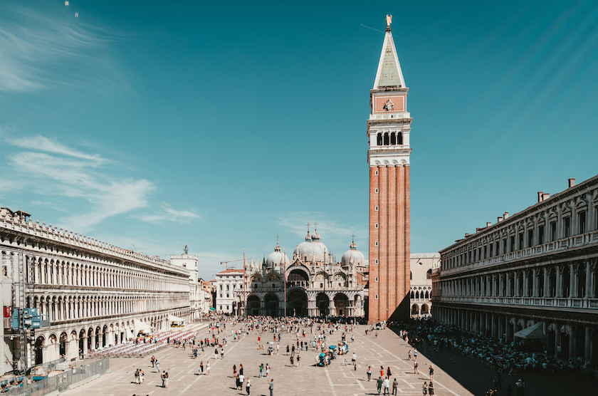 Un día soleado sobre una animada plaza histórica europea llena de gente, con un alto campanario de ladrillo y una gran basílica abovedada.