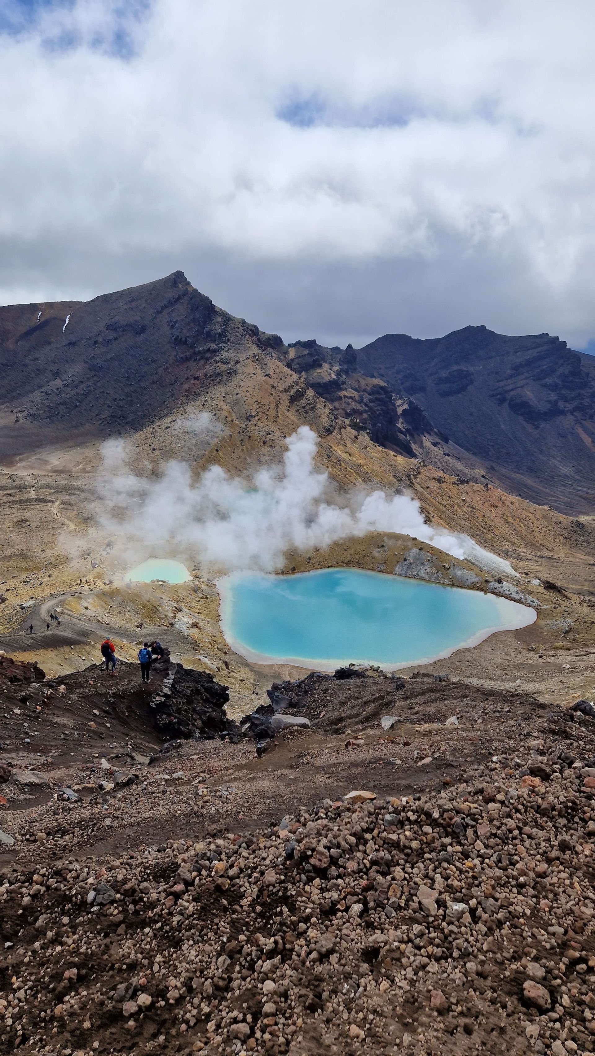 Un voyage de groupe WeRoad de randonneurs sur un sentier rocheux, surplombant deux lacs géothermiques bleus vifs d'où s'élève de la vapeur, dans une vallée volcanique.