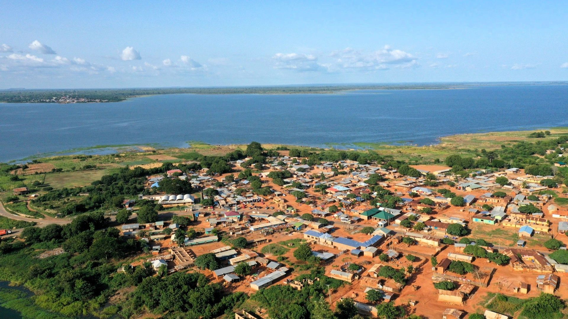 Luftaufnahme eines kleinen Dorfes mit unbefestigten Wegen und verstreuten Bäumen, gelegen am Ufer eines großen Gewässers unter blauem Himmel.