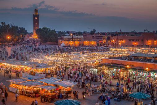 Una vista dall'alto di una piazza cittadina affollata al tramonto, piena di bancarelle illuminate e un alto minareto sullo sfondo.
