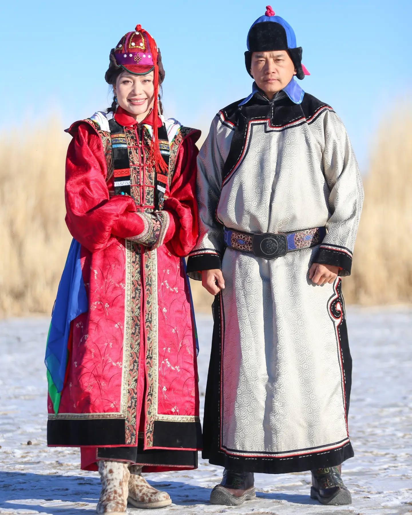 A man and a woman in colorful, traditional Mongolian attire stand side-by-side in a field under a clear blue sky.
