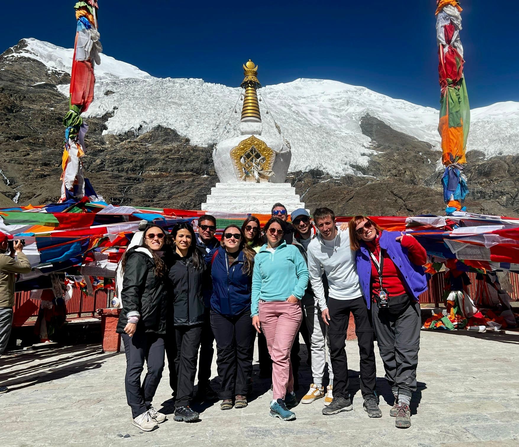 Un viaggio di gruppo WeRoad posa per una foto davanti a uno stupa bianco e bandiere di preghiera, con una montagna innevata sullo sfondo.