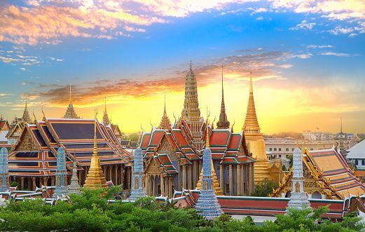 An ornate Thai temple complex with golden spires and tiled roofs against a colorful sunset sky.