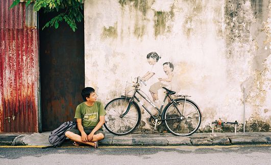 A young man sits on a sidewalk looking at a real bicycle parked in front of a mural of two children riding it.