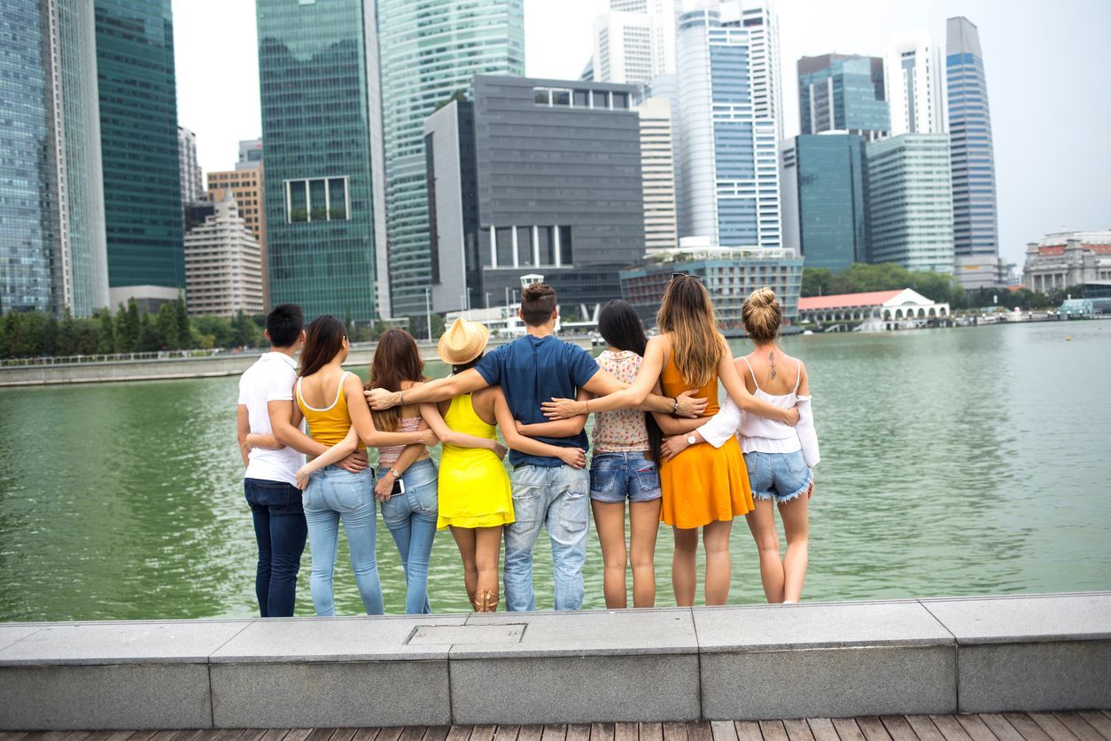 A WeRoad group trip seen from behind, standing with arms around each other looking out at a modern city skyline across the water.