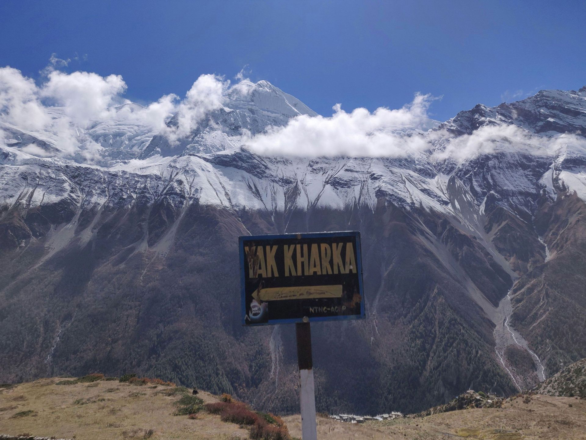 Un panneau "Yak Kharka" se dresse devant une vaste chaîne de montagnes enneigées sous un ciel bleu, avec un petit village dans la vallée en contrebas.