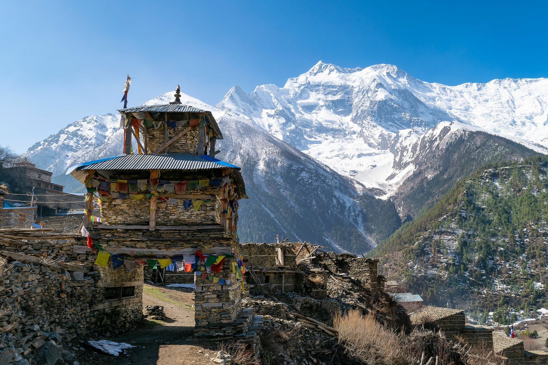Un portail en pierre orné de drapeaux de prière colorés se dresse dans un village de montagne, avec d'imposants sommets enneigés en arrière-plan.
