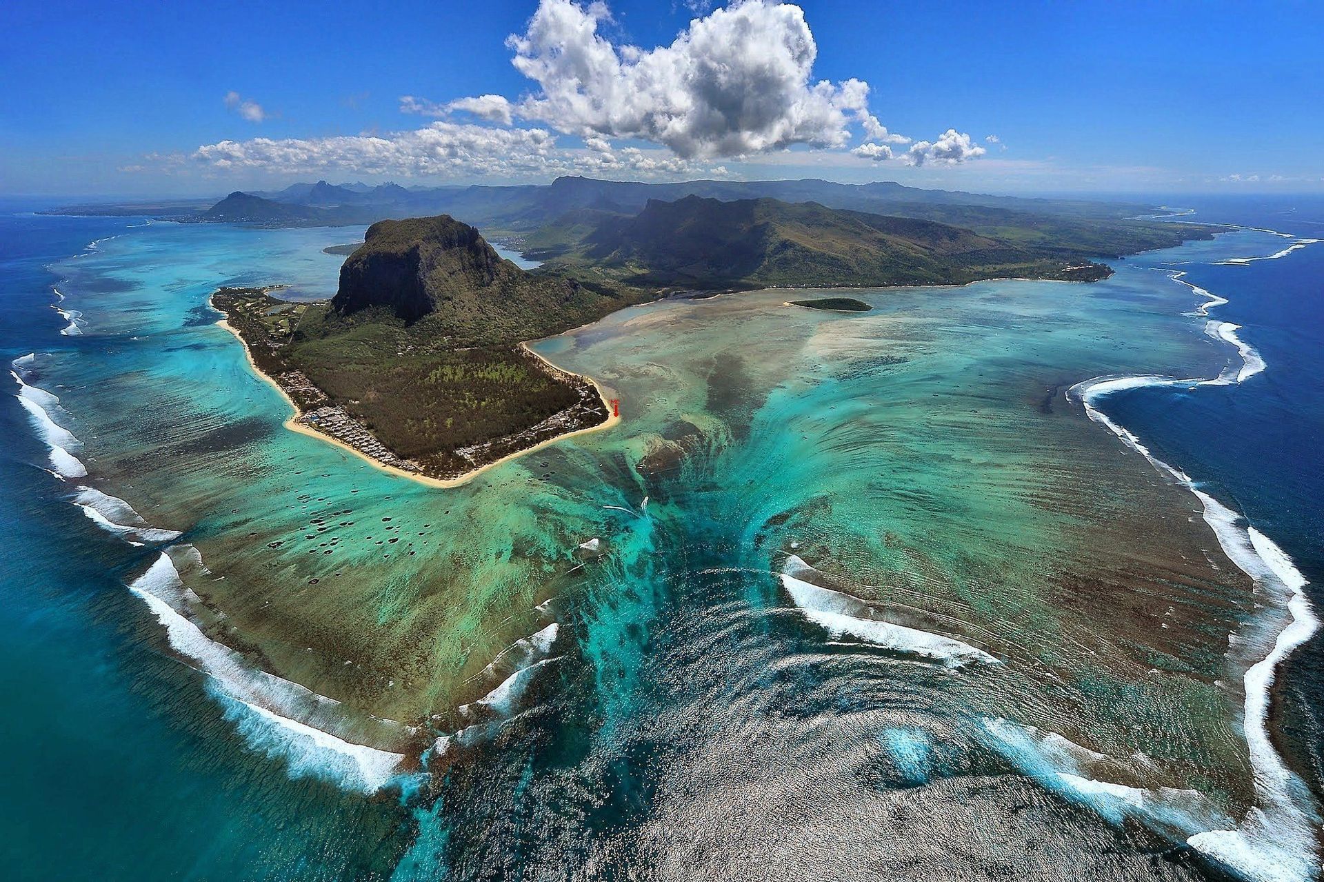 Vista aerea di una penisola tropicale, dove il movimento della sabbia crea l'illusione ottica di una cascata sottomarina nell'oceano turchese.