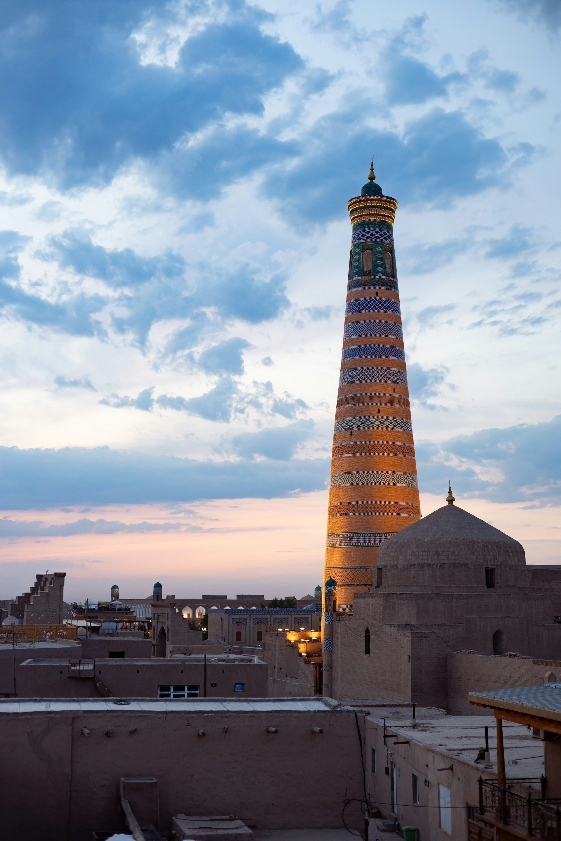 A tall, ornate minaret with blue and orange tiles rises above a cityscape of adobe buildings at sunset under a cloudy sky.