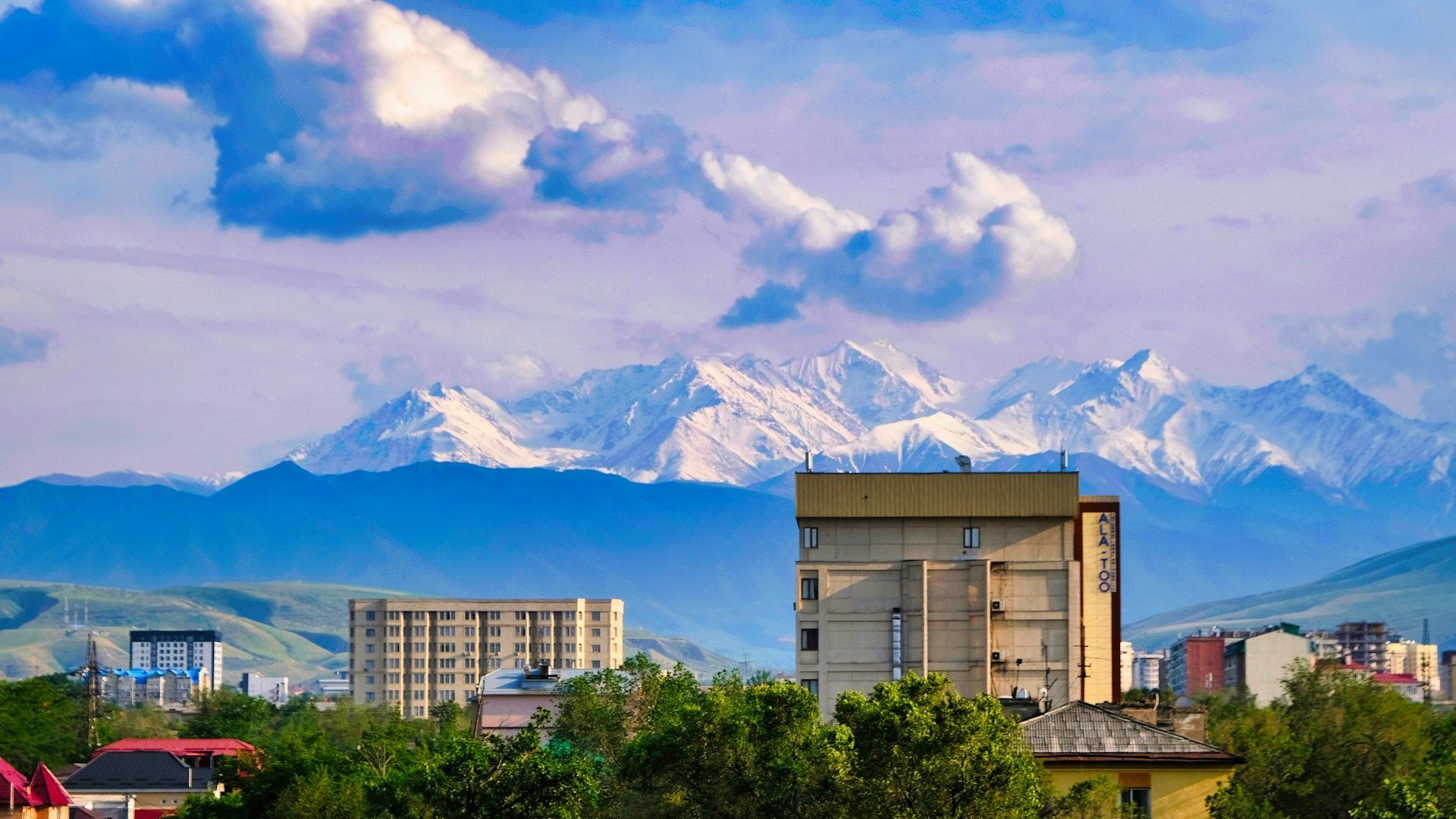 Uno skyline cittadino con edifici e alberi si trova di fronte a un'imponente catena montuosa innevata sotto un cielo azzurro nuvoloso.