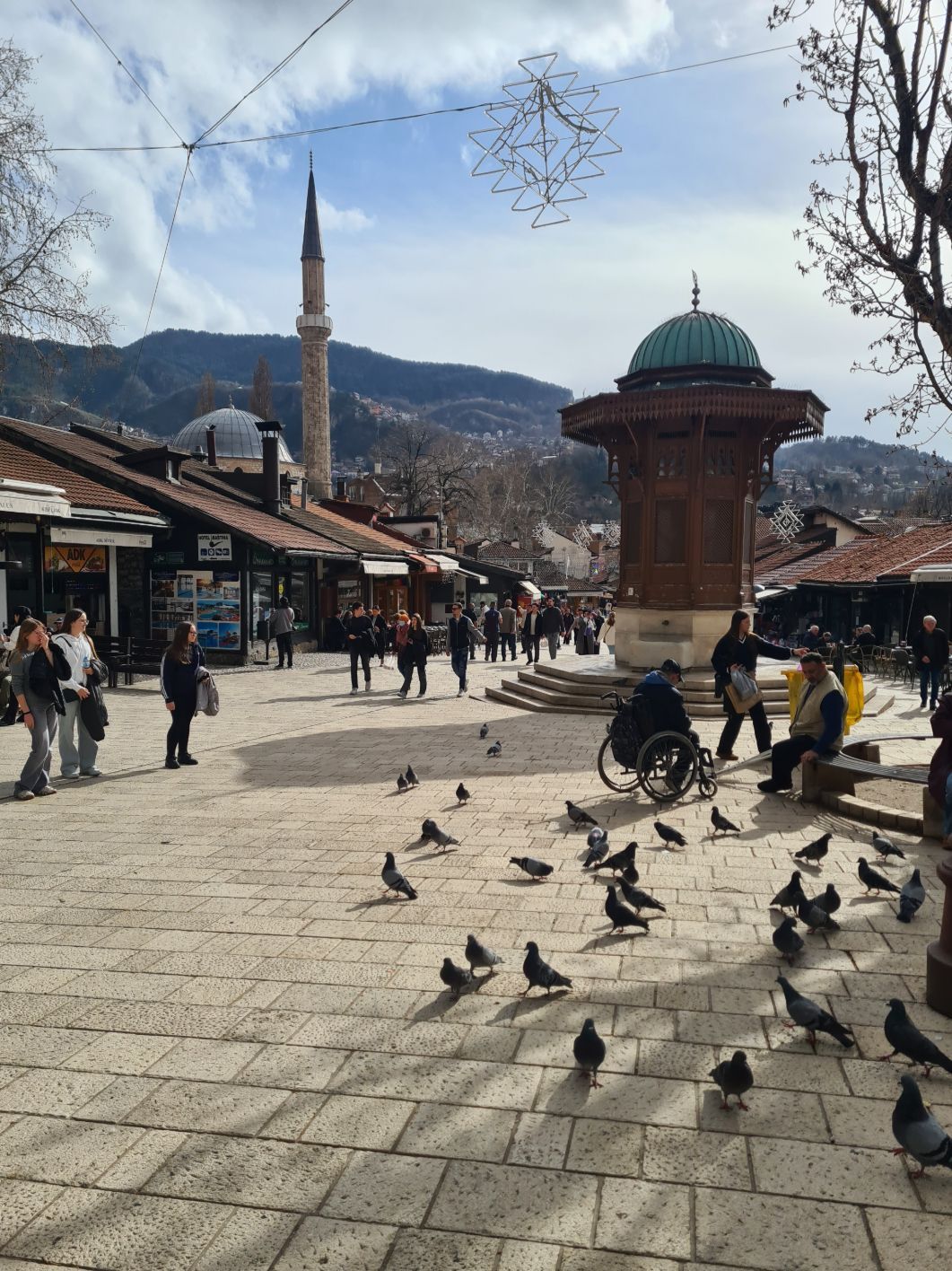 Menschen und Tauben versammeln sich auf einem historischen Stadtplatz mit einem markanten Holzbrunnen, einem Minarett und Bergen im Hintergrund.