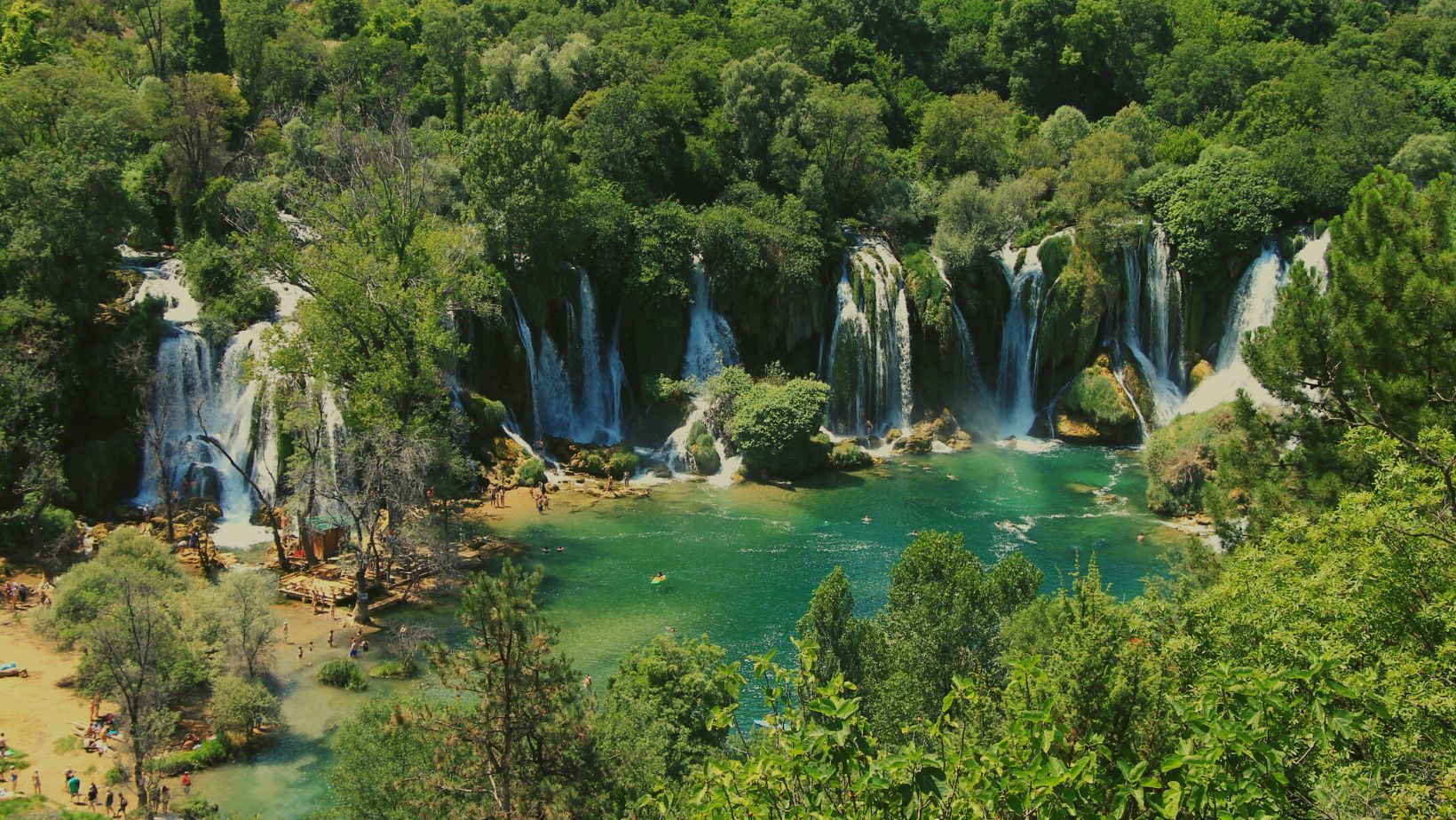 Breite Wasserfälle stürzen in einen türkisfarbenen Pool, in dem Menschen schwimmen, umgeben von dichtem grünem Wald.