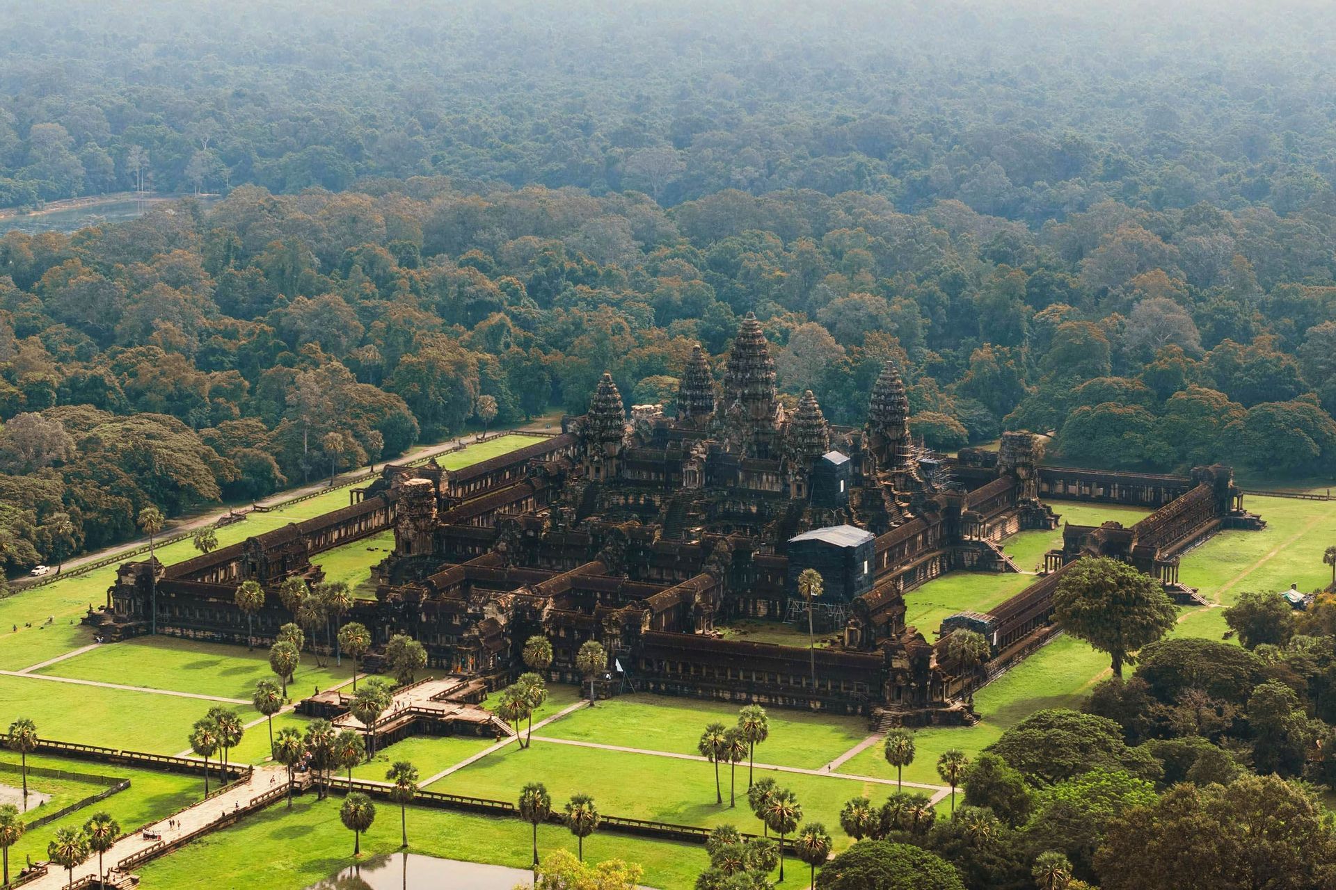 Una vista aerea di un grande e antico complesso di templi in pietra con torri, circondato da prati verdi, palme e una vasta giungla.