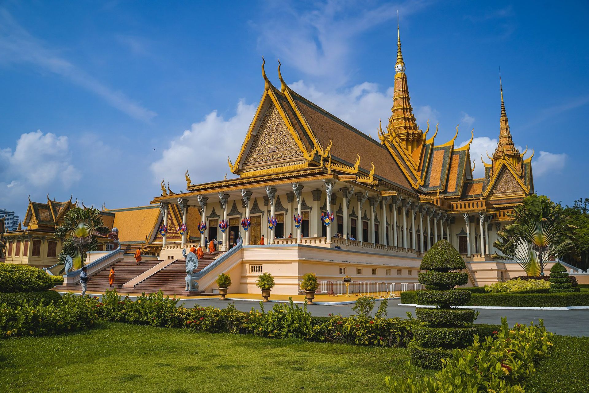 Monaci in tuniche arancioni salgono i gradini di un tempio ornato con tetti dorati, circondato da un prato verde sotto un cielo blu.
