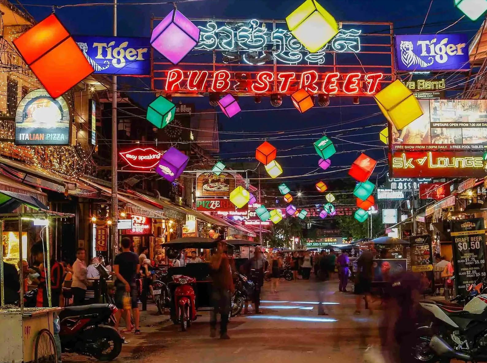 Una vivace strada notturna è illuminata da una grande insegna al neon 'Pub Street' e da colorate lanterne a forma di cubo appese in alto.