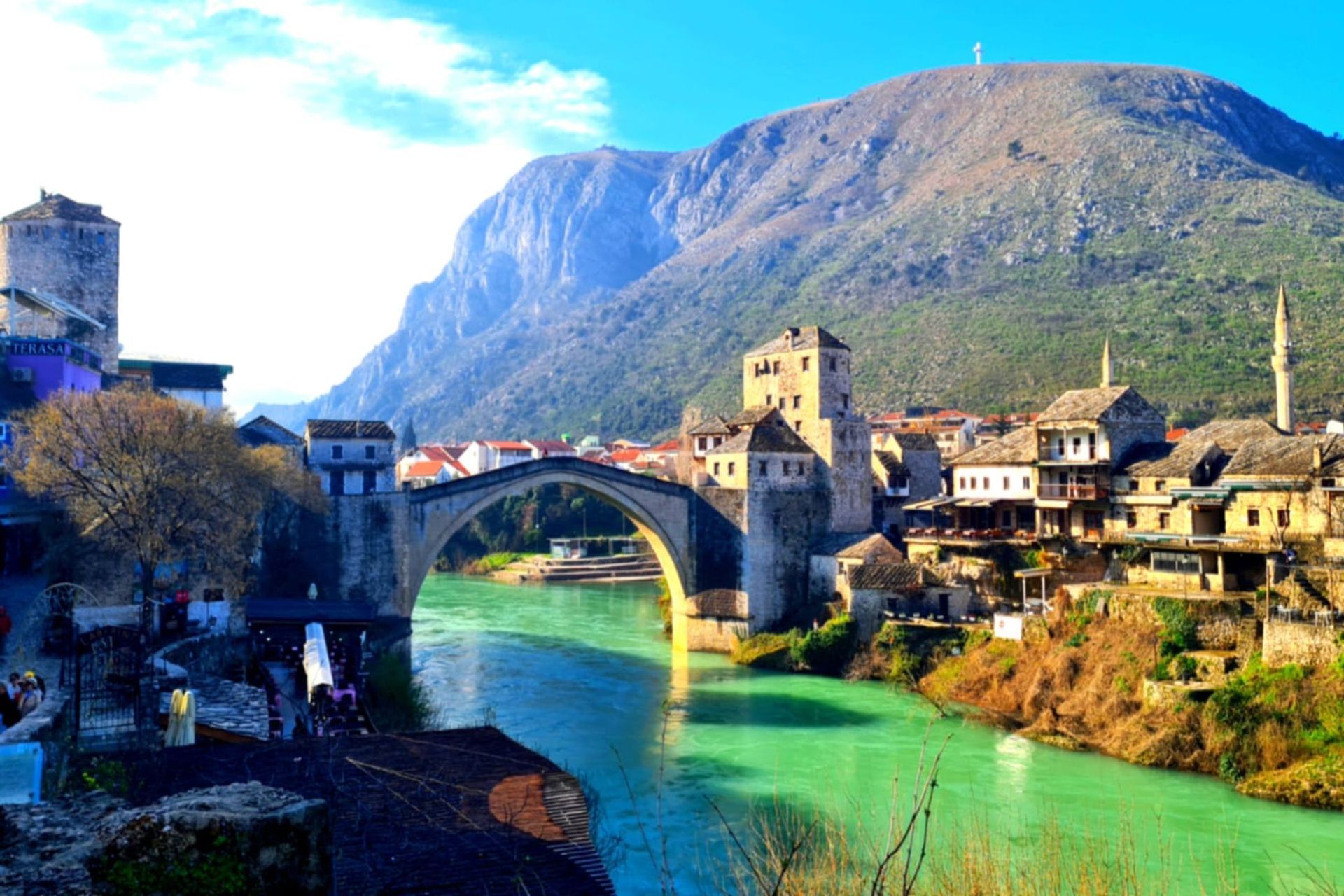 Eine historische Steinbogenbrücke überquert einen türkisfarbenen Fluss, der durch eine Altstadt am Fuße eines großen Berges unter blauem Himmel fließt.