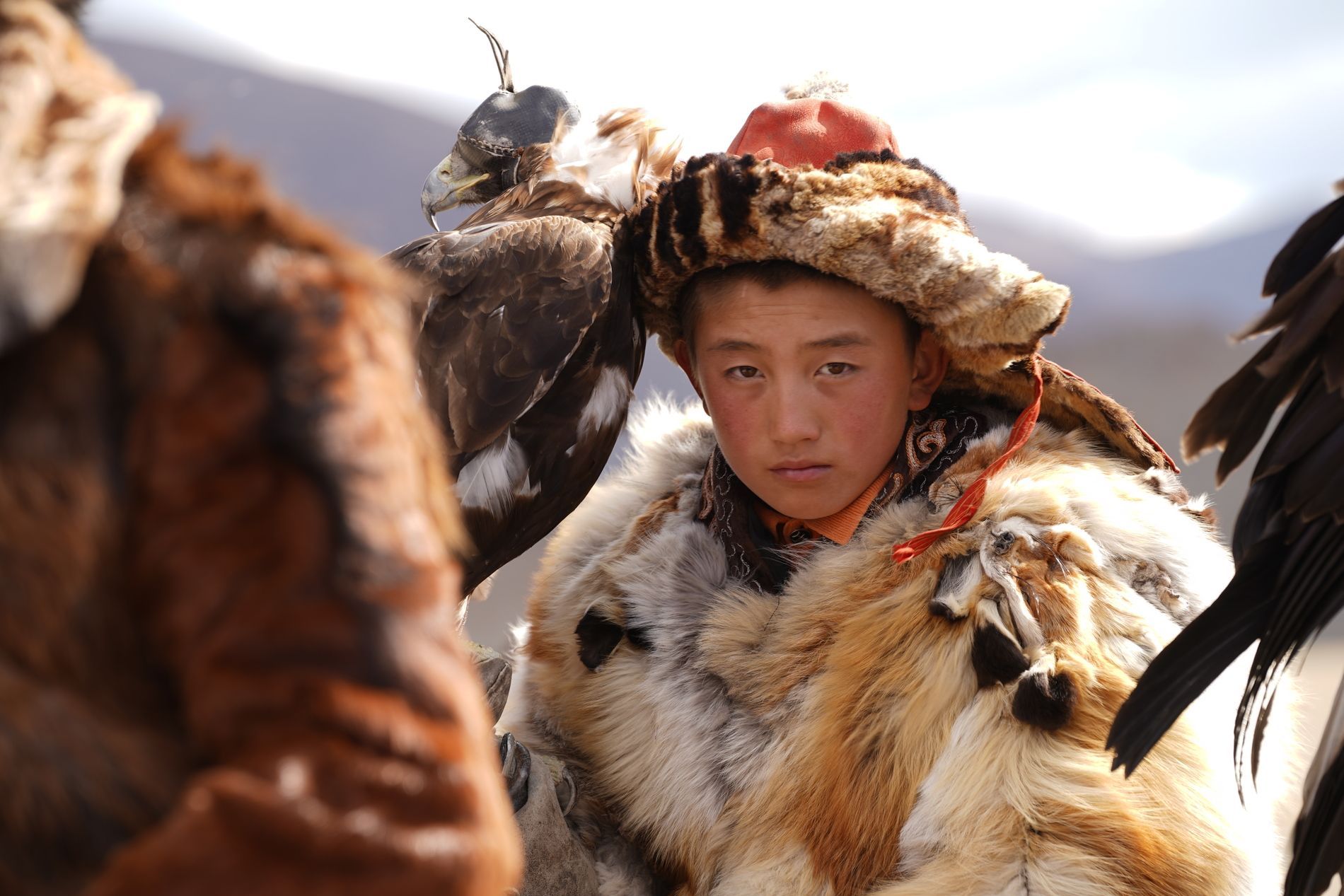 A young boy in traditional fur clothing and hat stands next to a hooded golden eagle, looking directly at the camera.