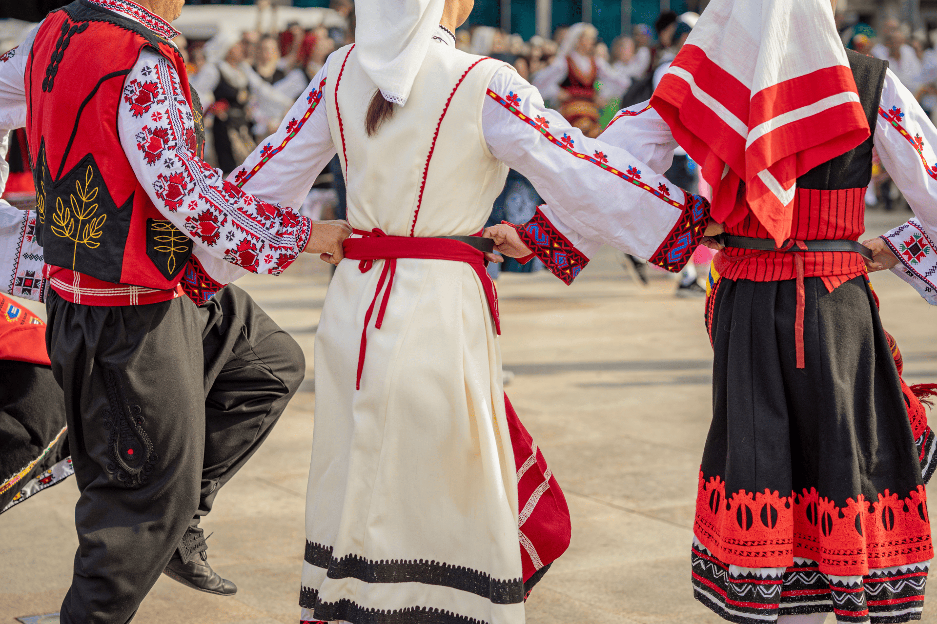 Tres personas vistas de espaldas, con trajes folclóricos bordados tradicionales, bailando en línea y sujetándose de los cinturones.